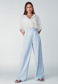 Lichtblauwe wijde broek met een hoge taille, knoopsluiting en twee voorkruimelzakken. Gecombineerd met een loszittend wit overhemden met knopen.