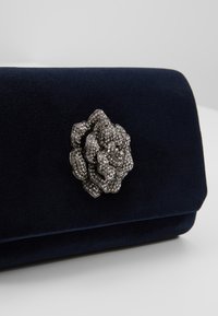 Pochette en velours bleu marine avec un fermoir en forme de rose incrusté de strass argentés centré sur le rabat avant.