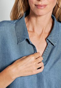 Helles blaues Denim-Hemd mit einem subtilen Glanz, featuring einen Kragen und eine partielle Knopfleiste. Die Hand des Models passt den Kragen an.