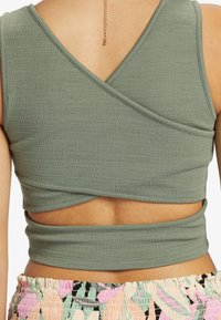 Vihreä oliivinvärinen kääntötoppi, joka on valmistettu teksturoidusta materiaalista. Siinä on ristiselkätyyli ja istuva vyötärö, jossa on aukko selässä.