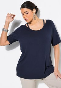 Marinblå t-shirt med kort ärm, rundad halsringning och lager av spaghetti-band. Mjuk tyg med avslappnad passform, i kombination med beiga byxor.