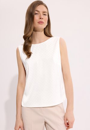 Kobieta z długimi falowanymi włosami, ubrana w białą, bezrękawnikową bluzkę z delikatnym wzorem w kropki oraz beżowe spodnie, na jednolitym jasnym tle.