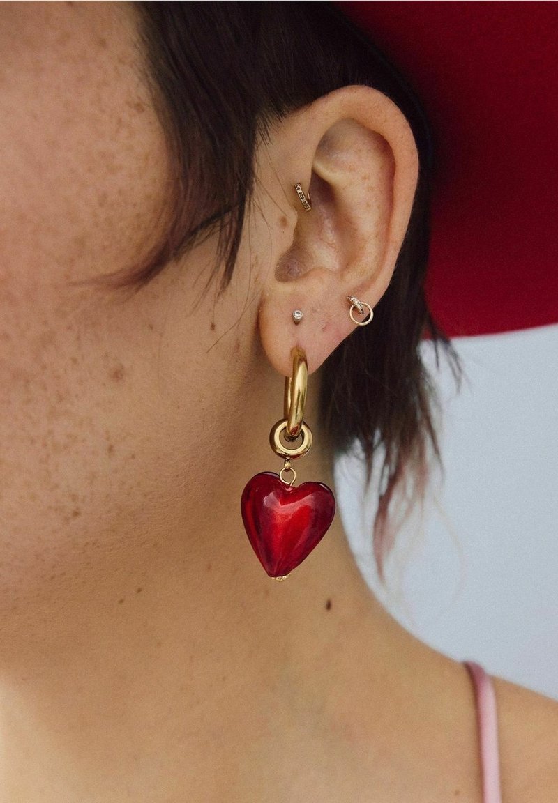 Gros plan d'une oreille avec trois petites boucles d'oreilles en or et un grand anneau en or tenant un pendentif rouge en forme de cœur.