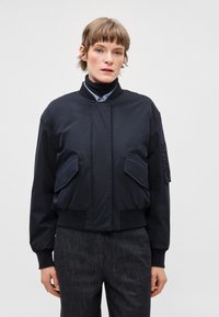 Tumši zila bomber stila jaka ar stāvošu apkakli, priekšējām atloku kabatām un ribotiem aprocēm. Gluda, matēta auduma, ar īsu piegriezumu.