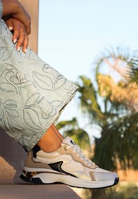 Sneaker beige e crema con dettagli neri, parte superiore texture e hardware dorato. Abbinato a pantaloni in denim ricamati di un blu chiaro.