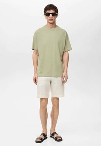 Svijetlozeleni kratki rukavi majica, bež kratke hlače i crne sandale. Glatka tkanina, opušteni kroj i jednostavan dizajn. Naočale za sunce dodaju modernu notu.