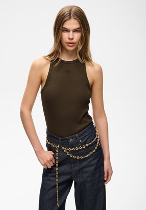 Jeune femme aux cheveux blonds et raides, portant un haut sans manches olive, un jean foncé et une ceinture chaîne dorée sur fond blanc.