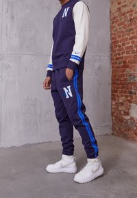 Marinblå och vit sweatshirt med blå ränder, prydd med en bokstav "N"-logotyp; matchande marinblå joggerbyxor med blå sidränder och logotyp. Vita sneakers.