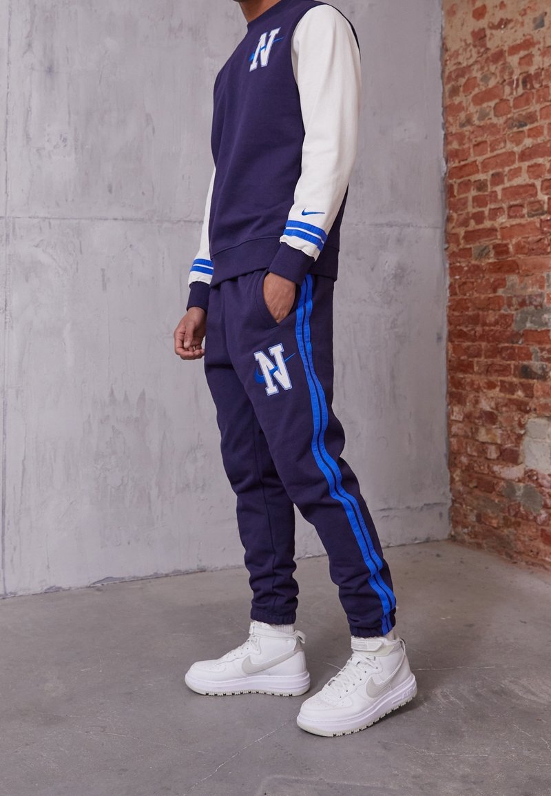 Marinblå och vit sweatshirt med blå ränder, prydd med en bokstav "N"-logotyp; matchande marinblå joggerbyxor med blå sidränder och logotyp. Vita sneakers.