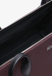 Sac à main bordeaux avec des accents en cuir noir, fermeture à glissière et intérieur lisse. Comprend des accessoires en métal et un détail de logo.