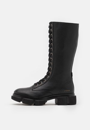Botte noire en cuir montant jusqu'au genou avec un bout arrondi, semelle en caoutchouc épaisse et œillets dorés, présentant un design à lacets sur le devant.
