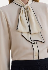 Cream blouse with a large, tied bow detail at the neck, featuring black piping along the edges. Smooth, lightweight fabric.