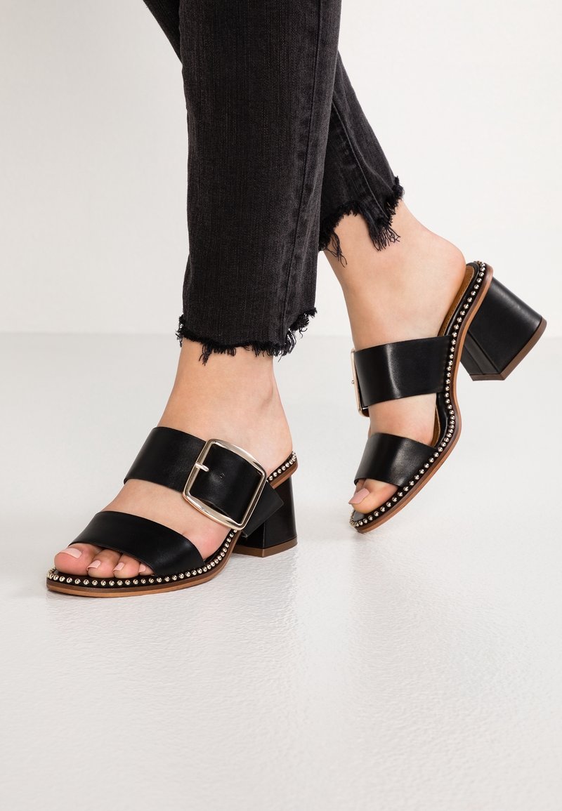 Pieds portant des sandales à talons blocs noirs avec de grandes boucles et des clous argentés le long de la semelle, associées à un jean skinny noir à ourlet effiloché.