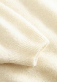 Kremo spalvos vilnonio megztinio audinys, rodantis švelnią tekstūrą, raštuotas rankogalių detalės ir smulkūs, pūkuoti pluoštai.