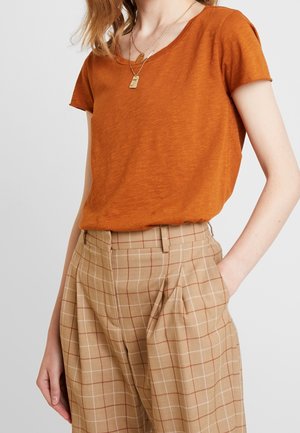Vrouw die een roestoranje korte mouwen top draagt, ingestopt in een high-waisted beige geruite broek met hand in de zak en meerdere gouden kettingen om.