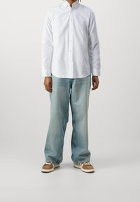 Camicia bianca a maniche lunghe con bottoni e orlo arrotondato, abbinata a jeans oversize blu chiaro e sneakers marroni con suole bianche e dettagli in bianco.