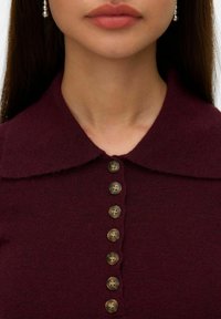 Mujer vistiendo un suéter burdeos abotonado con un amplio cuello y pendientes de perlas, mostrada desde el cuello hasta la parte superior del pecho.