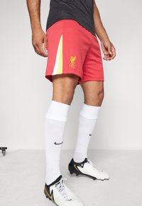 Punase Liverpool FC lühikesed püksid, millel on kollased aktsendid ja logo, koos pikkade valgete sokkide ja must-valgete jalgpalliketsidega. Sile kangast tekstuur.