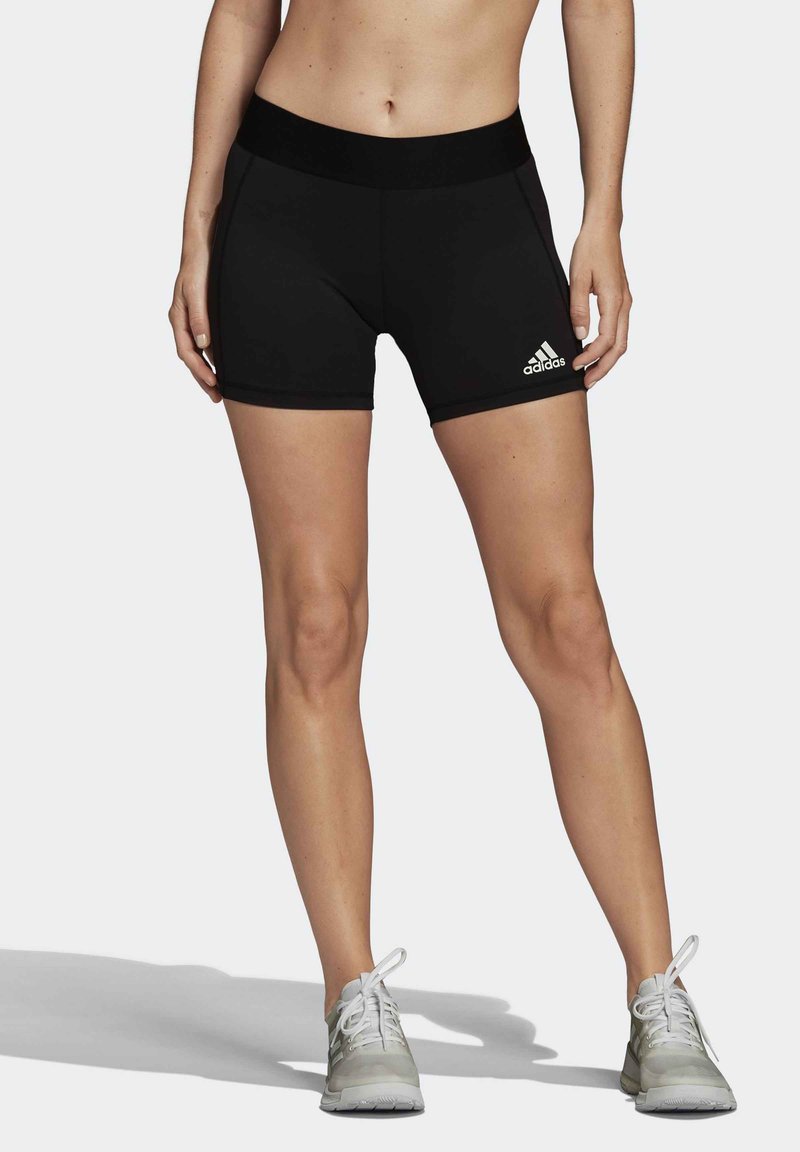 Shorts deportivos negros con una cinturilla ancha, hechos de un material elástico; presentan el logo blanco de Adidas en el lado izquierdo. Zapatillas plateadas completan el look.