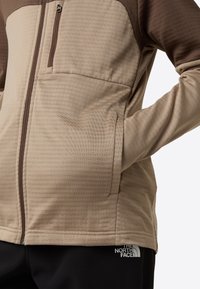 Jachetă bej cu fermoar, cu un model texturat, în dungi; are un buzunar pe piept și buzunare laterale; accente maro pe mâneci și guler.