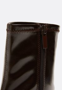 Gros plan sur une botte marron foncé brillante avec des coutures en zigzag visibles en haut et une fermeture éclair latérale fermée.