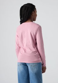 Žena s pletenicama koja nosi ružičastu majicu dugih rukava i plave traperice, stoji okrenuta leđima prema jednostavnoj sivoj pozadini.