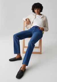 Vrouw met krullend haar zittend op een houten stoel, gekleed in een wit overhemd, licht jasje, blauwe spijkerbroek en zwarte loafers tegen een effen achtergrond.