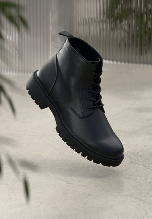Bottine noire en cuir à lacets avec semelle épaisse à crampons flottant au-dessus d'un sol en béton craquelé gris clair.