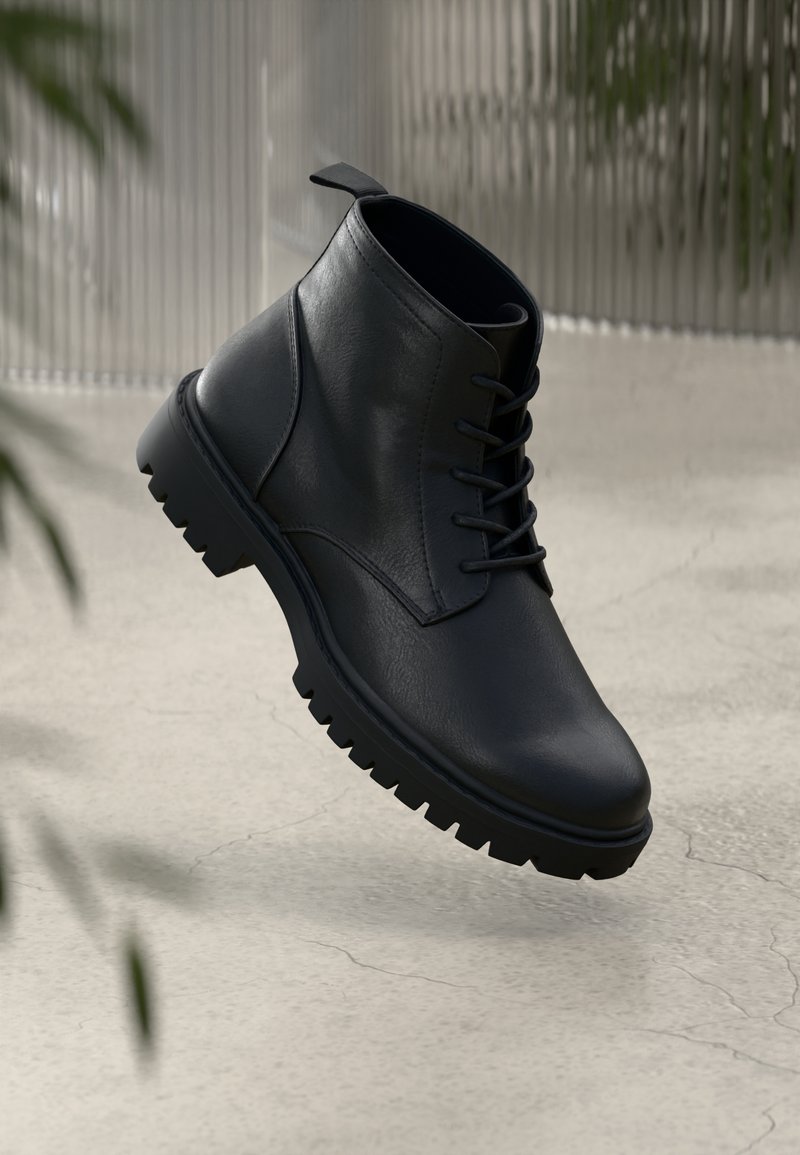 Bottine noire en cuir à lacets avec semelle épaisse à crampons flottant au-dessus d'un sol en béton craquelé gris clair.