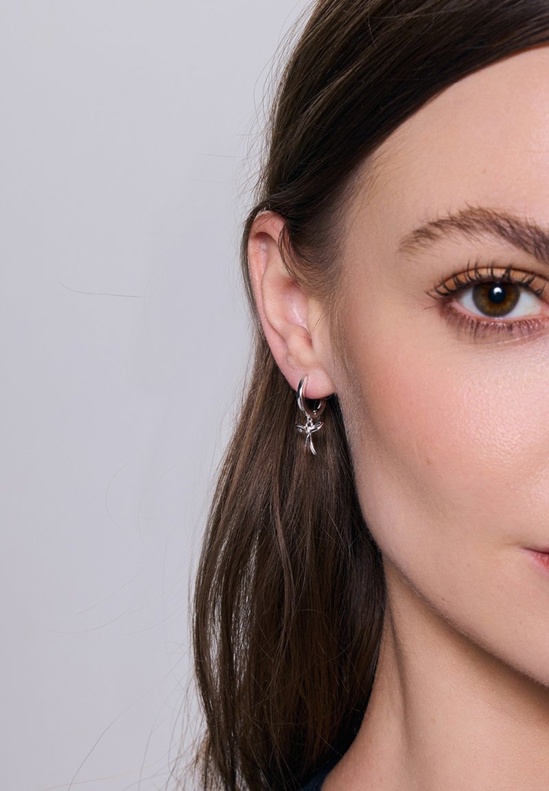 Srebrne kolczyki w kształcie kół z wiszącym akcentem w formie kwiatu. Kolczyki mają błyszczące wykończenie i kompaktowy design. Kolor skóry jest neutralny.