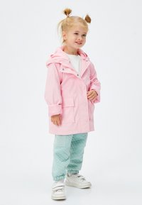 Chaqueta impermeable de color rosa claro con capucha, botones frontales y grandes bolsillos, combinada con pantalones verdes menta texturizados y zapatillas blancas.