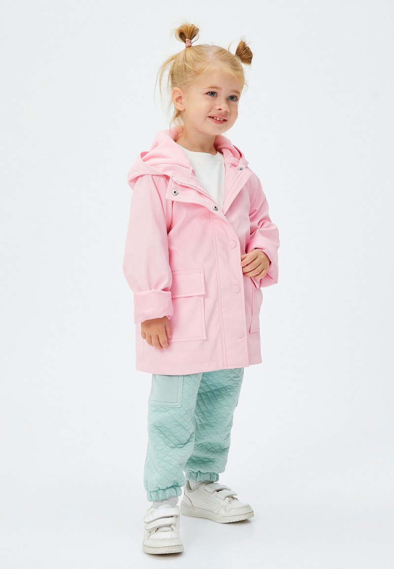 Chaqueta impermeable de color rosa claro con capucha, botones frontales y grandes bolsillos, combinada con pantalones verdes menta texturizados y zapatillas blancas.