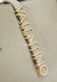 Beige läderväska med präglat "VALENTINO"-mönster. Har en logotyp i guldfärg och en texturerad rem. Slät yta.