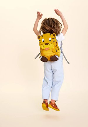 Gelber Rucksack mit Tiger-Thema aus Stoff, mit einem 3D-Gesicht, braunen Akzenten und verstellbaren grauen Trägern. Kind trägt hellblaue Latzhose.