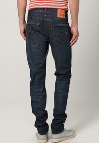 Vaqueros de mezclilla azul oscuro con un ajuste slim, que cuentan con un parche de cuero marrón, dos bolsillos traseros y detalles de costura sutiles.