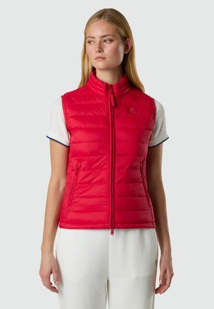 Donna che indossa un gilet imbottito rosso senza maniche, chiuso con una cerniera sopra una maglietta bianca a maniche corte, in piedi davanti a uno sfondo semplice.