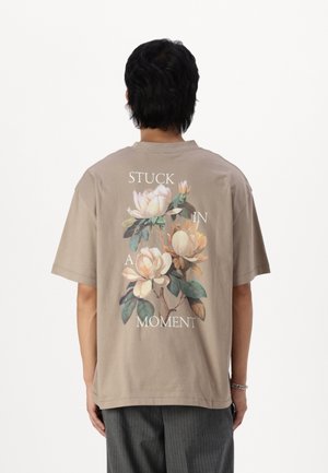 Osoba stojąca plecami do kamery, ubrana w beżowy T-shirt z kwiatowym wzorem i napisem "STUCK IN A MOMENT" białymi literami.