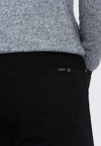 Pantalon en denim noir avec une texture lisse, arborant une petite étiquette en métal et des détails de poche arrière, associé à un pull en tricot gris clair.