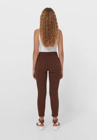 Jeans marrones ajustados con cintura alta y bolsillos traseros, combinados con zapatillas blancas. La tela parece suave y elástica.