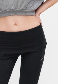 Μέση ενός ατόμου που φοράει μαύρα κολάν Nike και γκρι κοντό τοπ, με το ένα χέρι να στηρίζεται στο ισχίο.