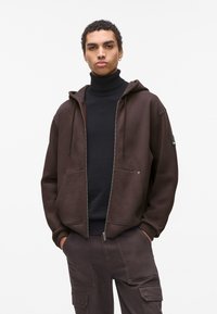 Rjava zip-up kapuca iz mehke tkanine, opremljena z kenguru žepom, nastavljivimi vrvicami in rebrastimi manžetami. Nošena čez črno turtleneck.