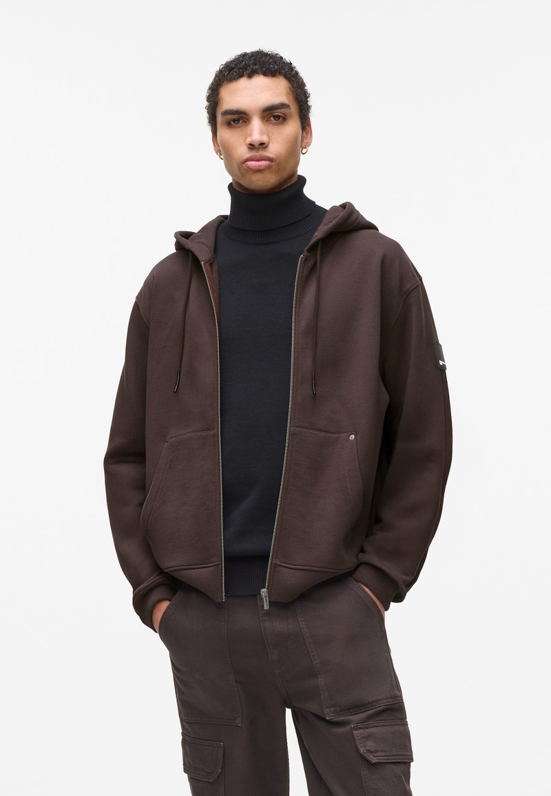 Rjava zip-up kapuca iz mehke tkanine, opremljena z kenguru žepom, nastavljivimi vrvicami in rebrastimi manžetami. Nošena čez črno turtleneck.
