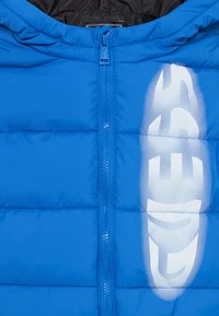 Giacca puffer blu con chiusura a zip, sezioni imbottite e un grande logo bianco nella parte anteriore. Texture liscia con una finitura lucida.