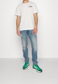 Man som bär en vit T-shirt med texten "LEE", ljusblå jeans och gröna och vita Adidas-sneakers som står mot en enkel vit bakgrund.