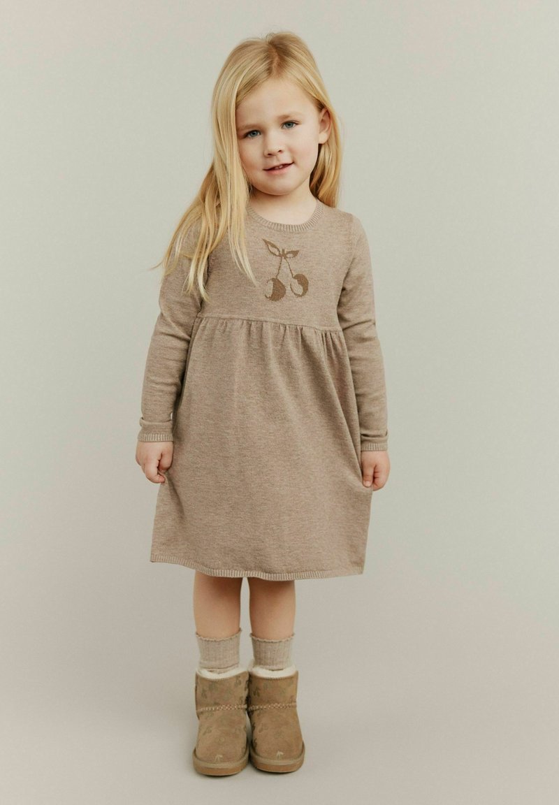 Smeđi pleteni kaput s dugim rukavima i nabrano strukiran, sa motivom trešnje. Uparen s bež čizmama obloženim krznom i svijetlim čarapama.