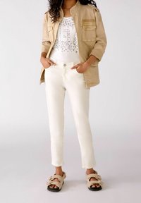 Beige jasje met twee voorzakken over een witte graphic tee. Witte slim fit broek en bruine fluffy sandalen maken de outfit compleet.