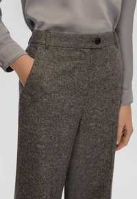 Donna che indossa pantaloni grigi in lana a vita alta con tasche laterali e chiusura a bottone, abbinati a una blusa grigia chiara.