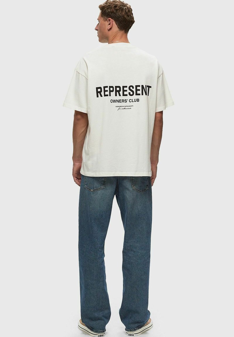 Camisa de algodón color crema con el texto en negrita negro "REPRESENT OWNERS' CLUB" en la parte de atrás. Corte holgado, mangas cortas, combinada con unos jeans azules.