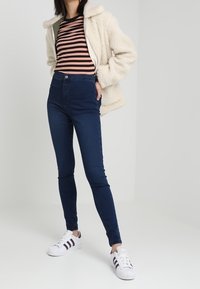 Kräppad krämfärgad jacka med fleecefoder, svart och rosa randig skjorta, mörkblå slim-fit jeans, vita sneakers med svarta ränder, stående pose.
