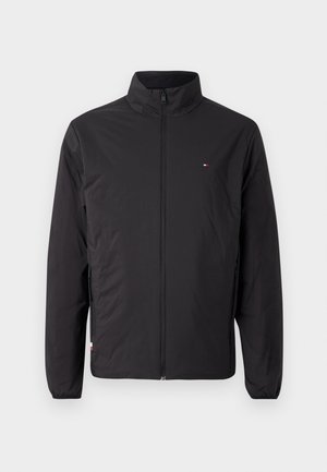 Musta kevyt takki korkealla kauluksella, täydellä vetoketjulla ja elastisilla hihansuilla. Omistaa pienen brändilogon rinnassa.